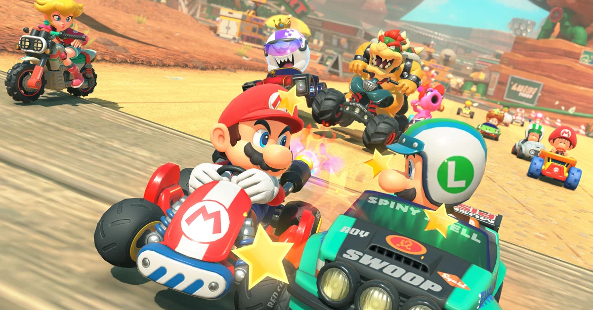 Mario and Luigi collide in a high-speed crash during a Mario Kart World race, with sparks and stars flying around them. Other characters, including Bowser, Princess Peach, Roy, and Birdo, race behind them on a desert-themed track, adding to the chaotic and action-packed scene.