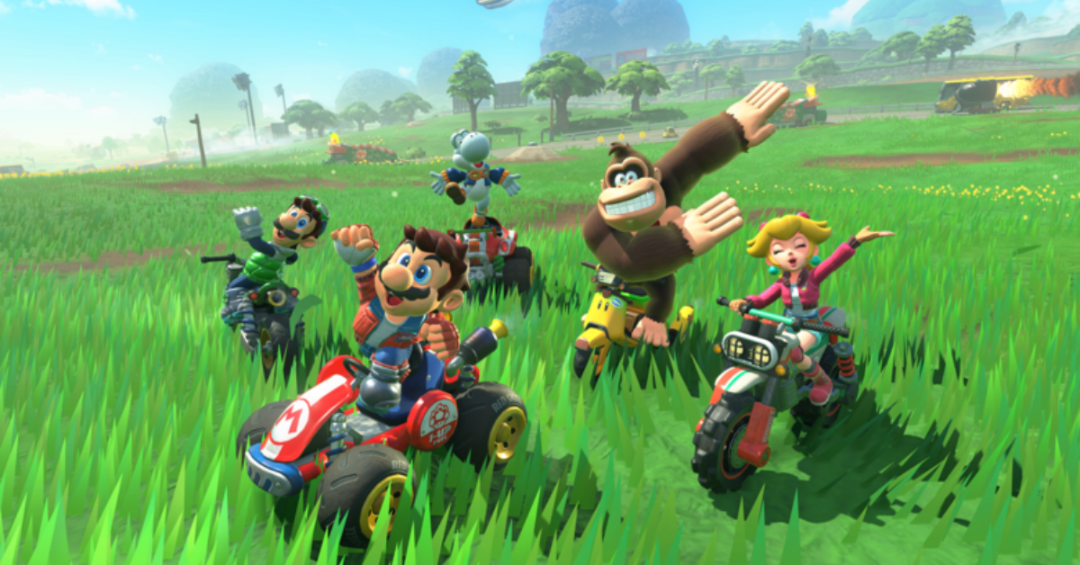Mario, Luigi, Yoshi, Donkey Kong, and Princess Peach celebrate cheerfully while riding their karts and bikes through a vibrant grassy field in Mario Kart World, with a scenic racetrack and trees in the background.
